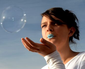 Production shot from 'Breathe', of a performer in teal lipstick blowing a bubble away from an outstretched hand.