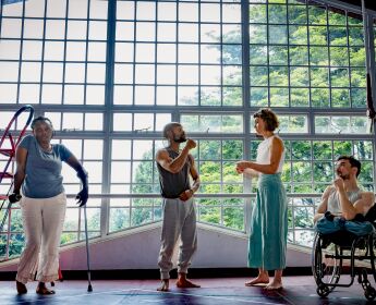 4 performers stand in by a large window of a studio in Kenya. Two performers stand, one is in a wheelchair and the other leans on crutches.