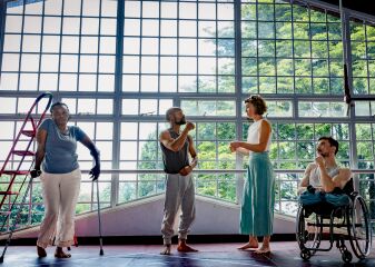 4 performers stand in by a large window of a studio in Kenya. Two performers stand, one is in a wheelchair and the other leans on crutches.