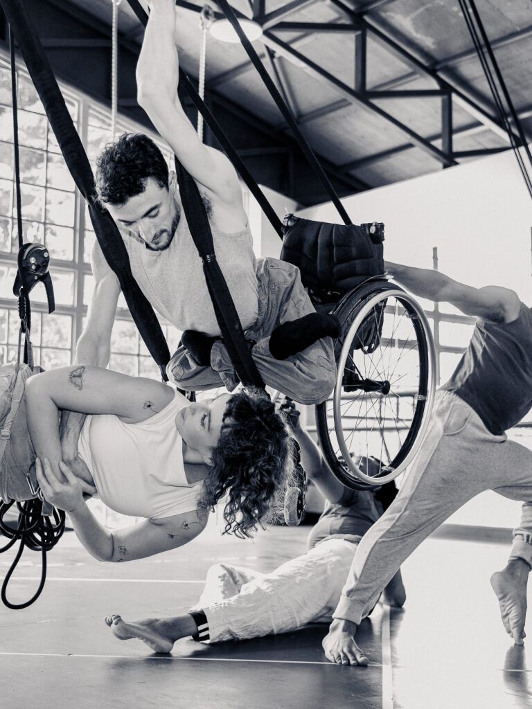 4 performers dance together. One is in a flying wheelchair, one is suspended in a harness. Two others are ground based dancers, interacting with those in the air.