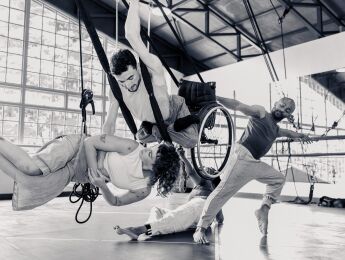 4 performers dance together. One is in a flying wheelchair, one is suspended in a harness. Two others are ground based dancers, interacting with those in the air.