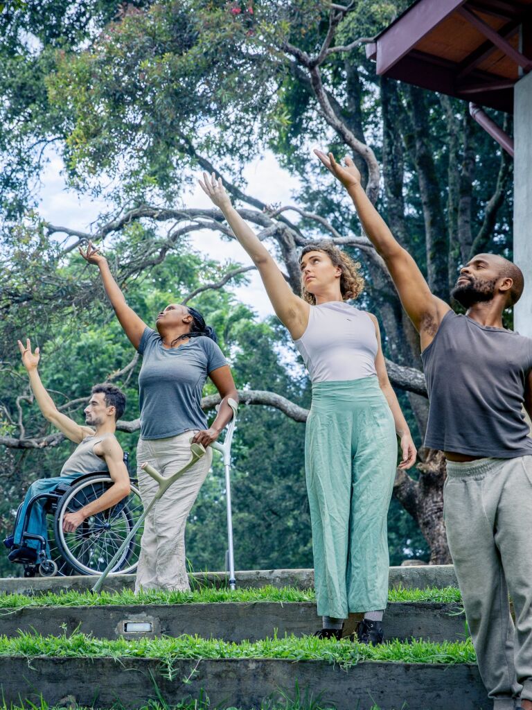 4 Performers stand in a line, all reaching their arm up. They are in a tropical garden, each on a garden step.
