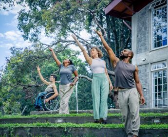 4 Performers stand in a line, all reaching their arm up. They are in a tropical garden, each on a garden step.