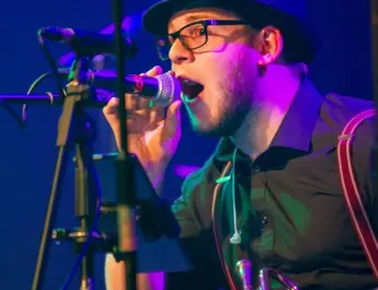 Harry, a young white man with a trilby hat on sings into the mic