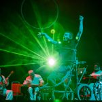 David Young moves across centre stage in his wheelchair walker, the Paraorchestra musicians seated behind him. David's arms reach up, it looks like he is catching the light from the bright green lasers coming from back of the stage.