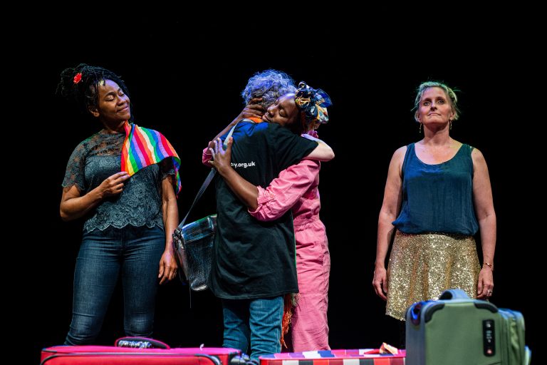 Four women standing in line. On the left, a black woman with a Pride rainbow flag on her shoulder smiles, she is looking at the two in the middle who are hugging. One of them wears a black Diverse City t-shirt and has grey curly hair and the other wears a pink one piece and has black skin. The fourth woman on the right looks in front of her with a proud and determined smile.