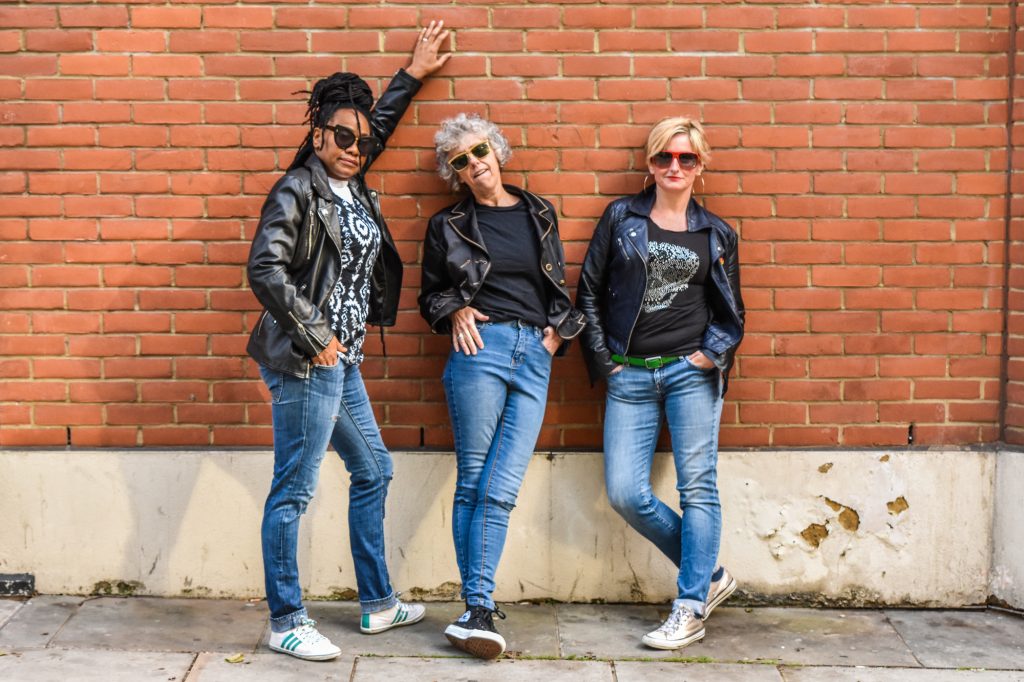 Three women in jeans and leather jackets, wearing sunglasses. They look cool. They pose to the camera leaning against a brick wall, with attitude.