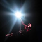 Performance of becoming: One member balances on a trapeze with legs pointed straight and arms raised above head. A bright light behind illuminates the performer.