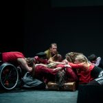 Performance of Becoming - EBYA members are gathered centre stage crouched and kneeling down in a circle. Everyone wears red/grey/black clothing.
