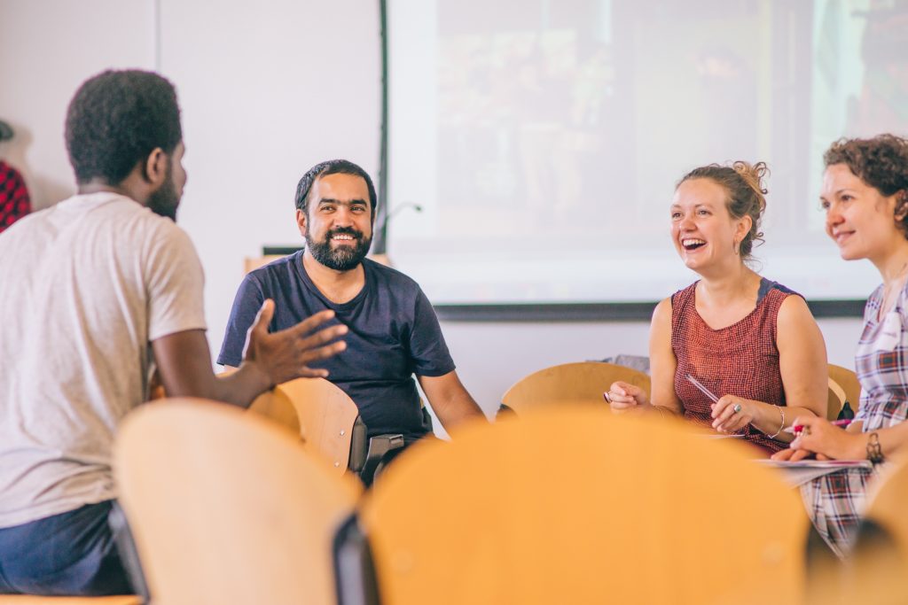 A small group of people sit in a room with lots of scattered chairs - it looks like a break from a workshop. They are talking and smiling together.