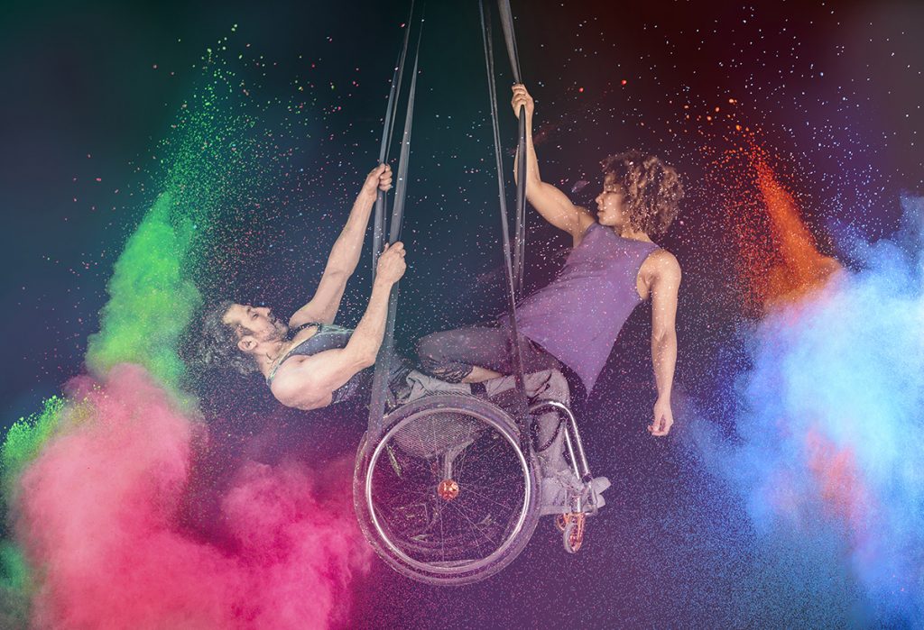 2 aerial artists suspended and entwined from straps: a white man in a wheelchair faces a muscular black woman - she is kneeling on his legs. There is an explosion of powdered paint around them in pink, green, blue and orange.