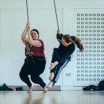 Two female workshop participants are suspended in harnesses, moving around above the floor.