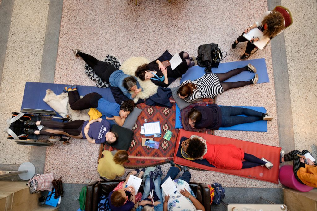 Photo from birds eye view - a group of 12 people in a circle, laying on the floor on mats and blankets, sitting on chairs and sofa. They are having a discussion; some people are taking notes on paper.