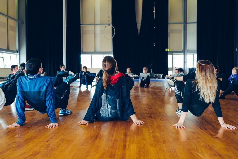 A group of workshop participants sat in a circle, with hands reaching behind their back, palms to the floor - one leg is ontop of the other knee. It looks like a good warm up stretch!