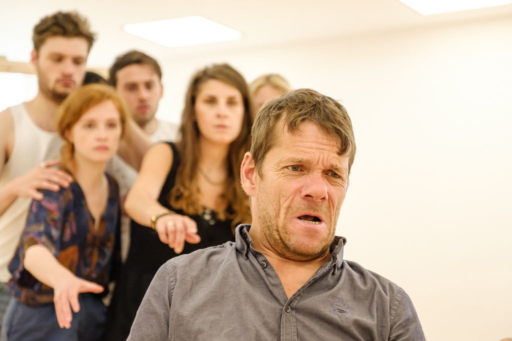 In the rehearsal room: Jamie is in the foreground, a distressed and concerned expression on his face. The actors gathered behind him are reaching forward, pointing at him with stern and serious facial expressions.