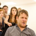 In the rehearsal room: Jamie is in the foreground, a distressed and concerned expression on his face. The actors gathered behind him are reaching forward, pointing at him with stern and serious facial expressions.