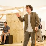 In the rehearsal room: Jamie sits in the background on a box, a BOV Theatre School student wearing a trilby hat and holding a wooden cane.