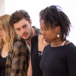In the rehearsal room: A white brunette actor, his head over other's shoulders, glares across the room at a black actress. The girls have a sad expression.