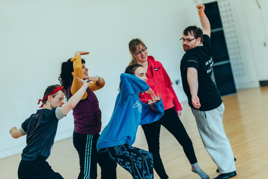 A group of 5 young workshop participants move around a studio space; they are lunging, stretching, twisting, reaching and smiling.