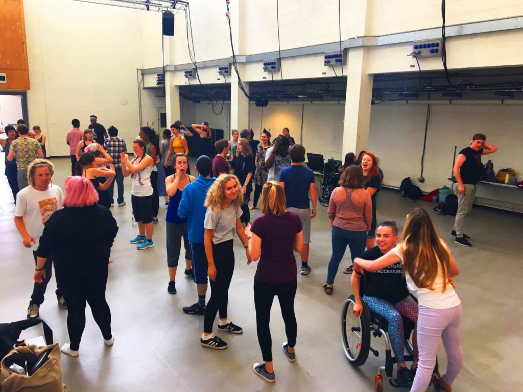 A large group of young artists from EBYA and National Youth Theatre gather in a large studio space. They are standing and exchanging conversations, everyone looks happy, laughing, smiling.