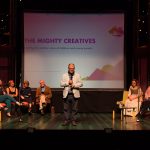 Nick Owen, standing centre stage, presentation behind reads 'The Mighty Creatives'. 12 others sit in a semi circle around Nick.