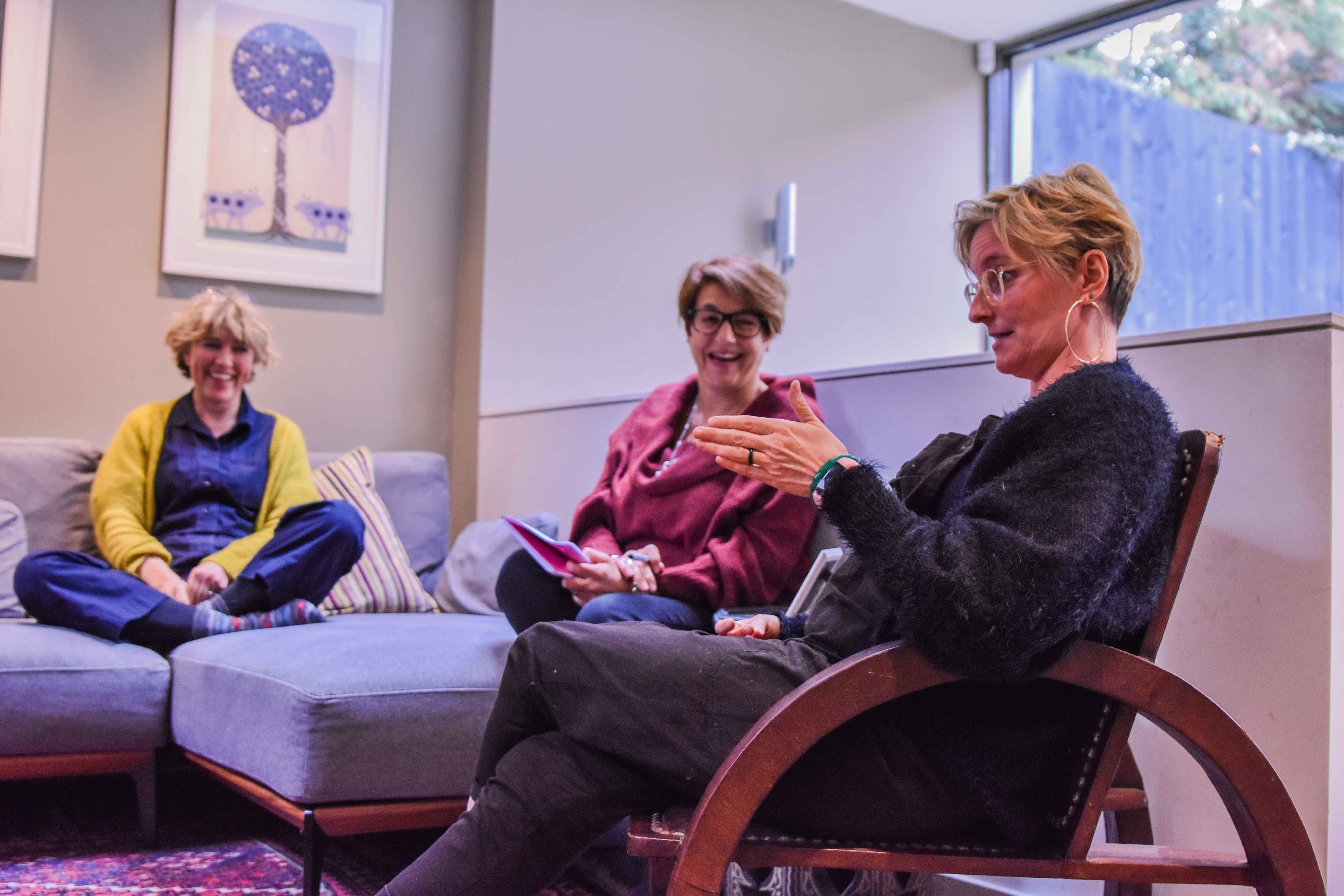 3 women in discussion in a living room, Claire Hodgson the show creator with her hands joined in support of what she is expressing while the other two are laughing
