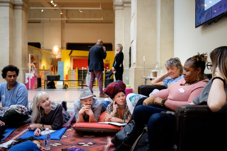 A diverse group of people gathered around sofa and rugs in the grand space at Bristol Museum. The group is deep in conversation.