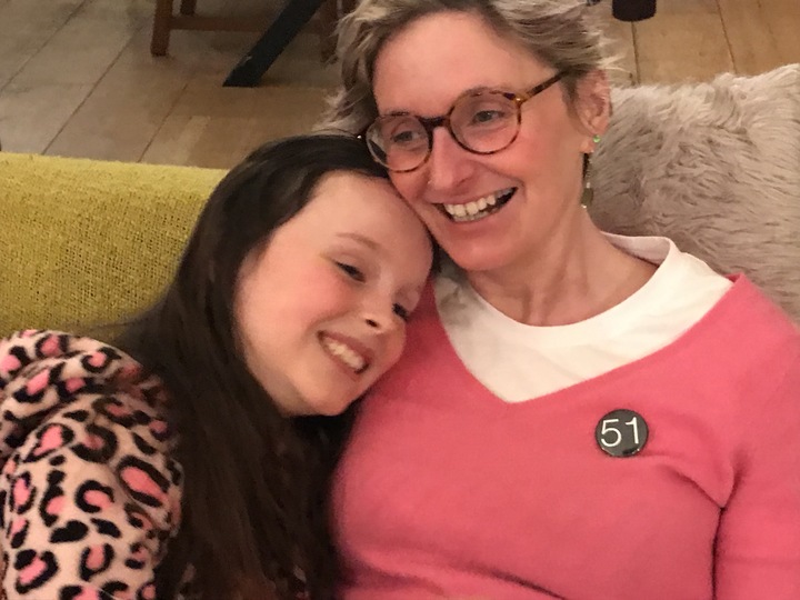 Claire Hodgson sits on the sofa with her young daughter, Scarlett. They are both smiling and hugging. Claire wears a badge that says '51' - representing 51% of the female population. 