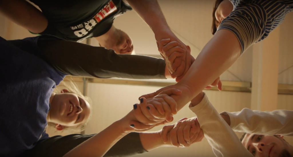 A group of young people clasp hands together - the camera is looking up between their hands to the ceiling - smiling faces are peeking between arms and hands.