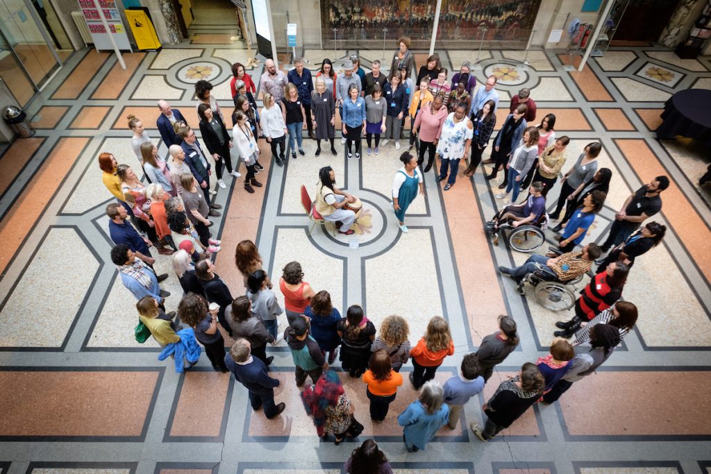 Doing Things Differently at Bristol Museum & Art Gallery. A diverse group of about 100 people are in a large circle in the grand entrance room to Bristol Museum. In the centre of the circle, a black man and black woman are leading the group through a movement exercise.