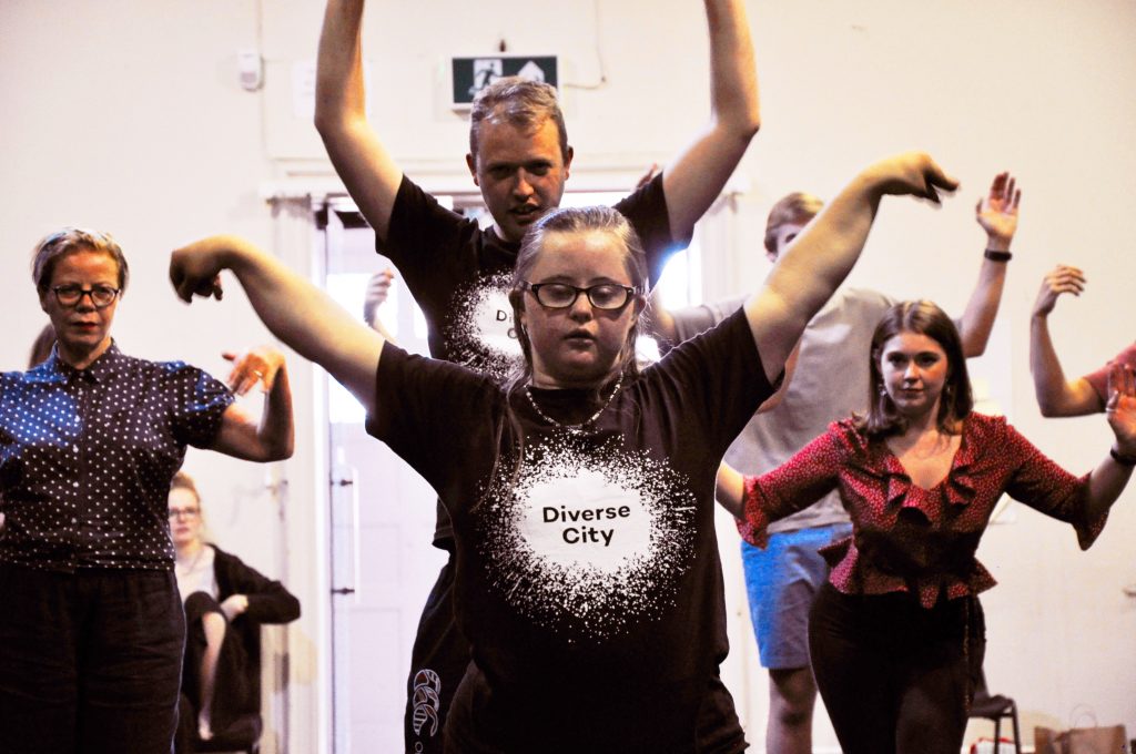 A young woman wearing glasses and a black t-shirt with a white Diverse City logo on it in the foreground. Her arms spread to her side, eyes closed. Behind her, several other performers making the same move.