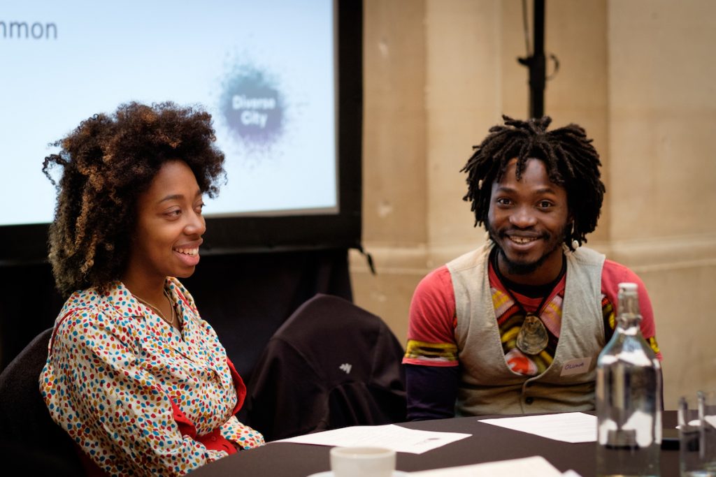 A young man of colour and a young woman of colour sit a table. In the background, a screen with a Diverse City logo