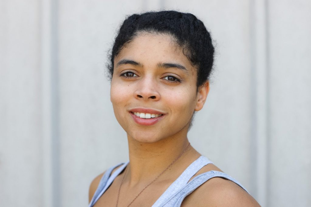 Portrait of a young woman of colour with dark hair tied back, brown eyes and a silver necklace. She smiles at the camera.