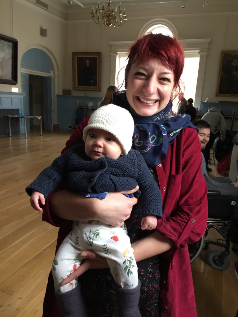 A smiling woman with red hair holds a baby with a white hat and dar blue jacket on inside an events venue.