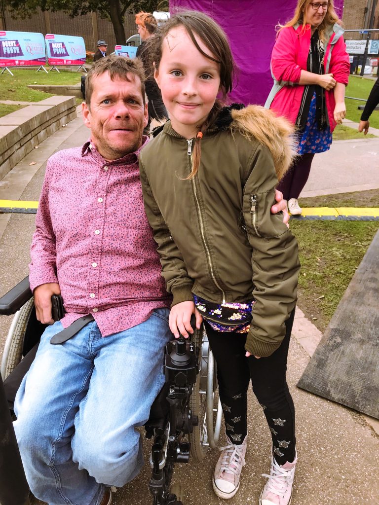 A man in wheelwhair with short brown hair and a red shirt and a young girl with long brown hair tied back and a kaki jacket pose together in an outdoors environment.