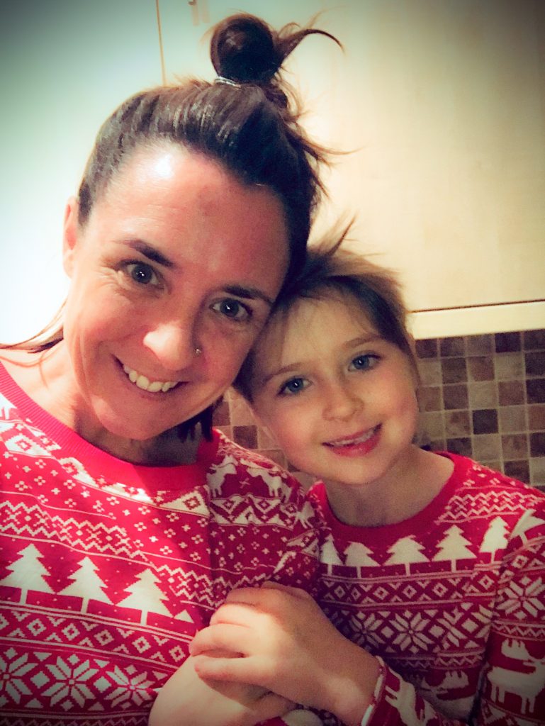 A woman with brown hair in a loose bun and a young girl with brown hair smile, both wearing red and white Christmas jumpers.