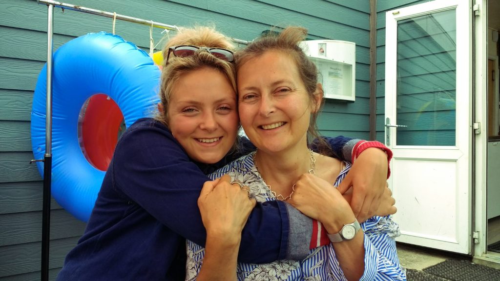 A young woman with blonde hair tied up and sunglasses propped up on her head holds and a woman who has long light brown hair and blue eyes. They hold each other around the shoulders and smile.