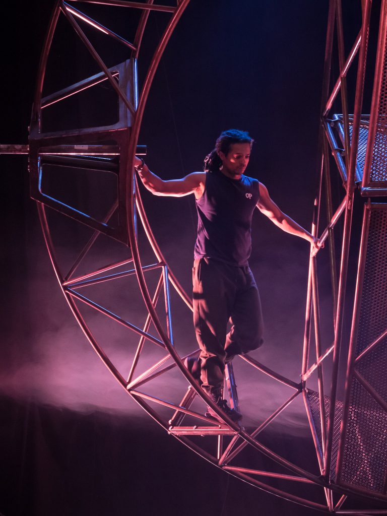 A man of colour with dreadlocks stands on a giant steel wheel, holding on to the bars.