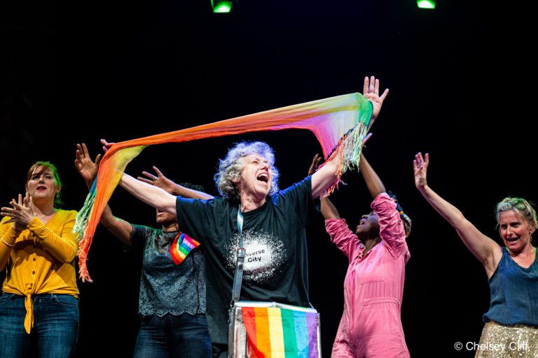 A middle-aged white woman waving a rainbow-coloured scarf above her head on a dark stage. Behind her, a black middle aged woman and a white middle aged woman, a young black woman, and a young white woman BSL interpreting. They all celebrate, hands up in the air.