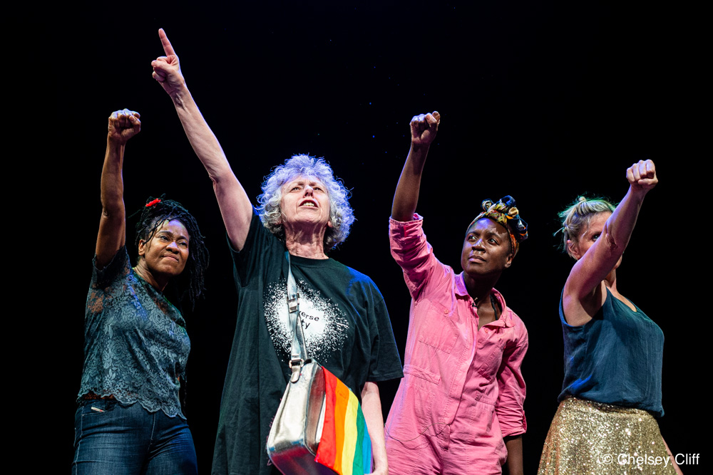 A middle-aged white woman raising her hand above her head on a dark stage, a rainbow-coloured satchel to her side. Behind her, a black middle aged woman and a white middle aged woman and a young black woman. They all hold their hands up in the air.