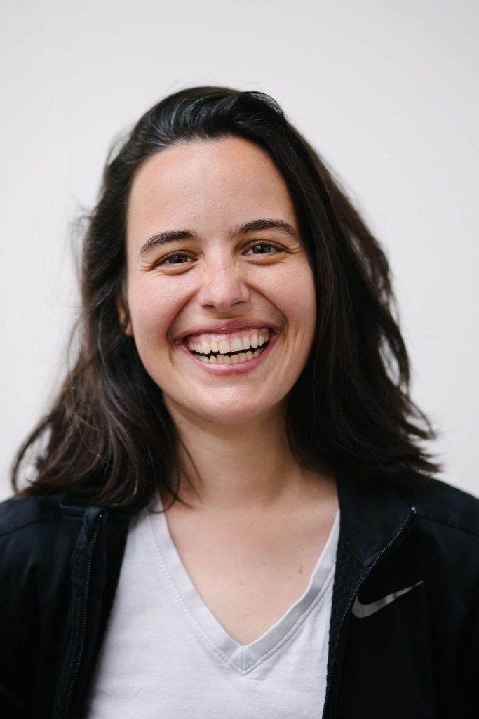 An olive skinned, dark haired woman smiles broadly - she is wearing a grey t-shirt under a black hoodie.