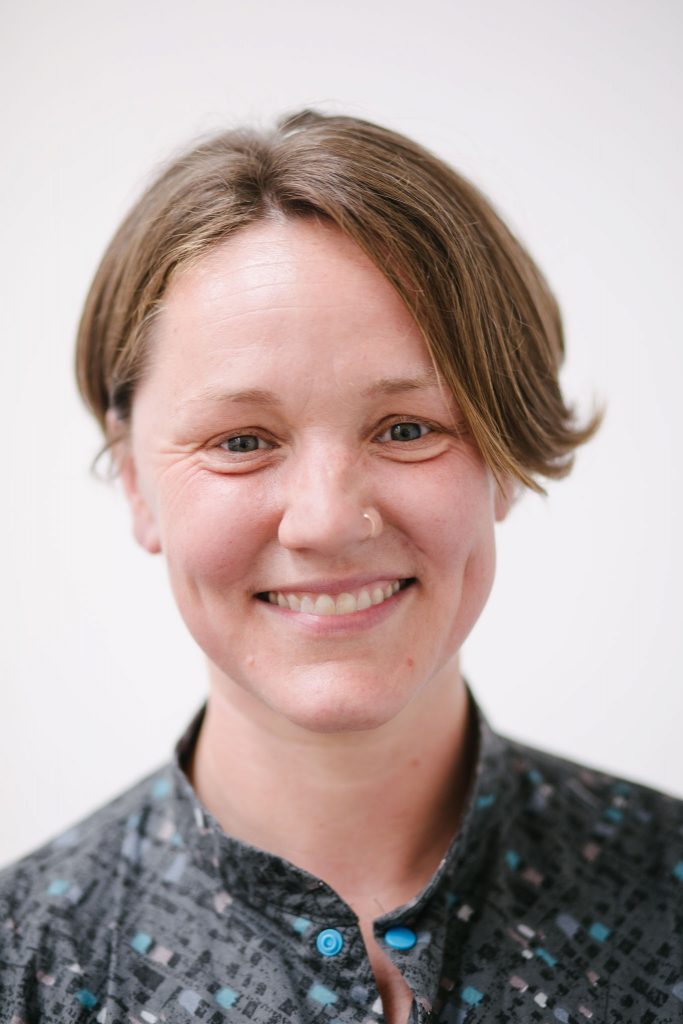 A smiling white woman with short, light brown hair, wearing a patterned jumpsuit.