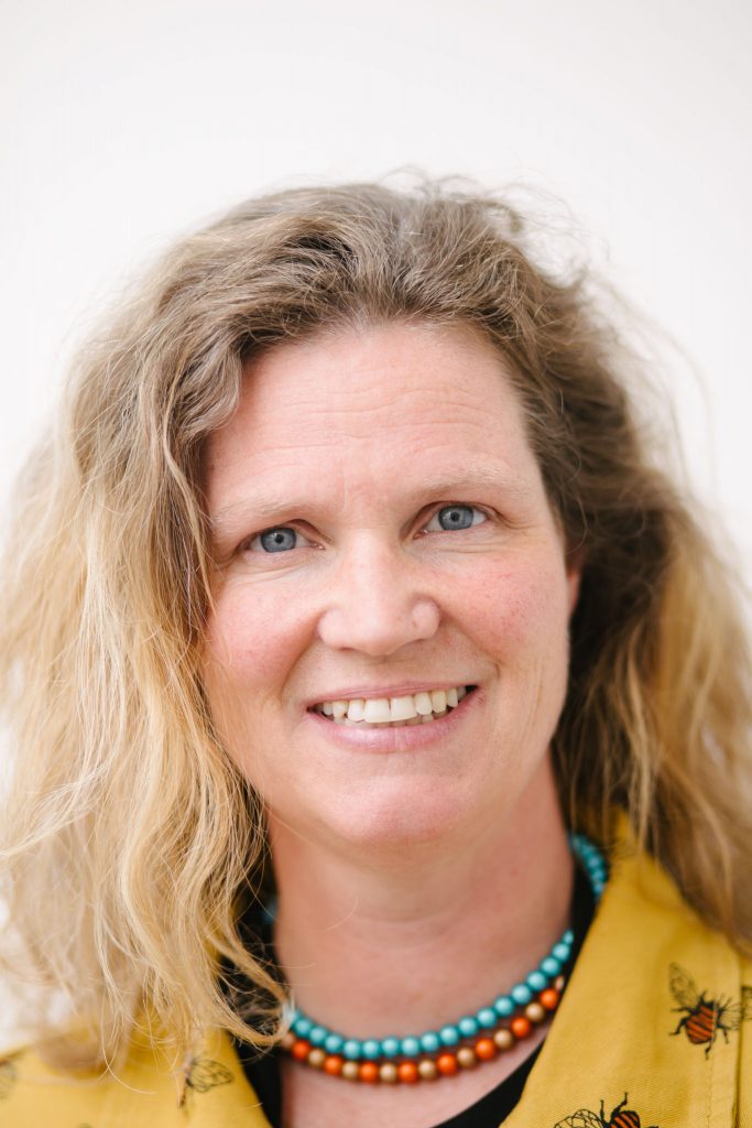 A smiling white woman with wavy fair hair. She wears a yellow jumpsuit with a bee pattern, and a blue and red necklace