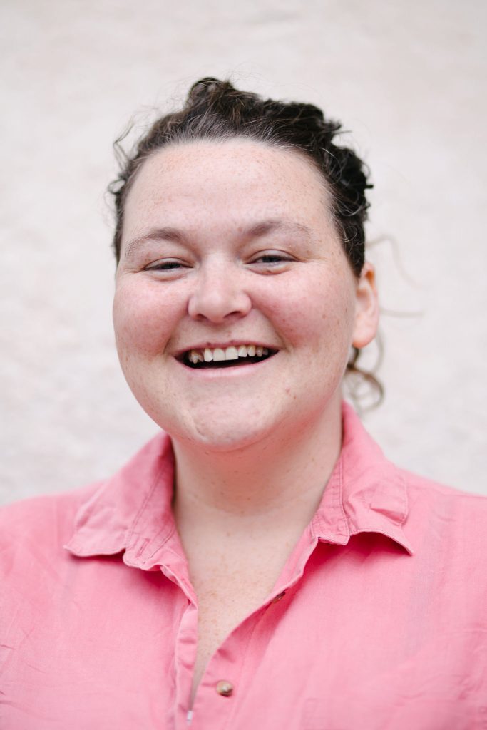 A smiling white woman with her dark curly hair tides back, smiles broadly. She is a wearing a pink shirt.