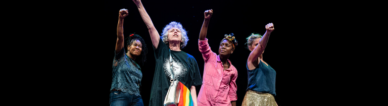 A middle-aged white woman raising her hand above her head on a dark stage, a rainbow-coloured satchel to her side. Behind her, a black middle aged woman and a white middle aged woman and a young black woman. They all hold their hands up in the air.