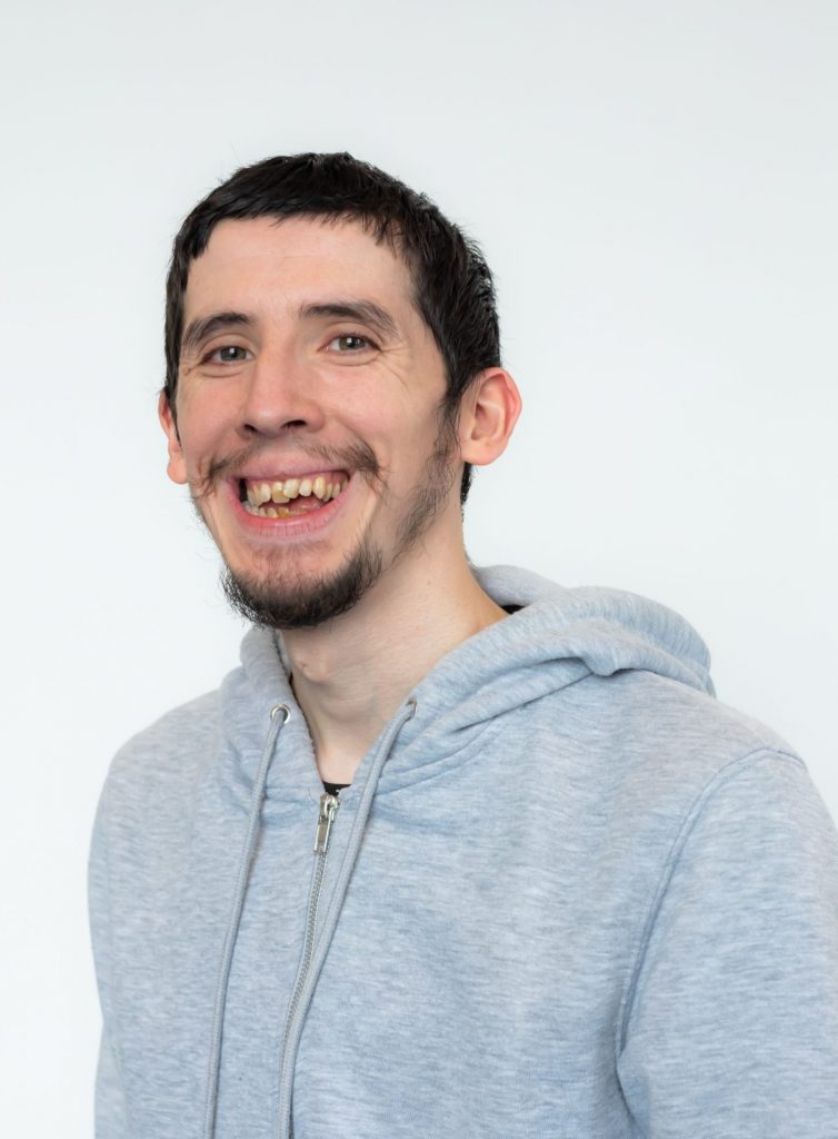 A white young man with dark hair and a goatee, wearing a grey hoodie. he smiles at the camera.