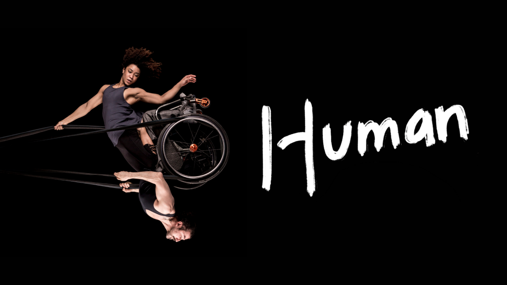 Black background. A photo of a woman and a man swinging in the air like a pendulum, looking powerful and graceful. They share a wheelchair which is held up in the air by black circus straps. She has frizzy brown hair, muscular arms and wears a grey tank top She is kneeling on his thighs and one of her arms holds the straps above her head while the other points down towards the floor. He has a neatly trimmed brown beard, muscular arms and wears a black tank top. He holds the straps to the sides of his chest with both hands. The image captures them at the highest point of the swing, to our right, bringing the man to an upside- down position with the woman above him. Their bodies are doing something hard, but their strength makes it look effortless. One word written in white, thick, brush-style letters above the photo: 'Human'.