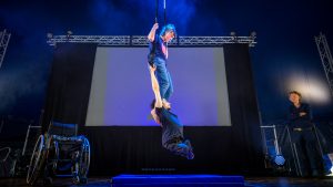 A woman is sitting on a trapeze, and hanging below her, a young man is hanging by his arms.