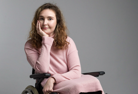 Meg Fozzard, a white woman with curly long, fair hair sits with her face cupped in her chin in her wheelchair. She wears a soft pink dress, black tights and ankle boots.
