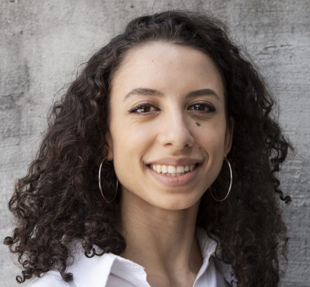 A woman with light brown skin and dark curly hair smiles at the camera. She is wearing a white shirt and hoop earrings